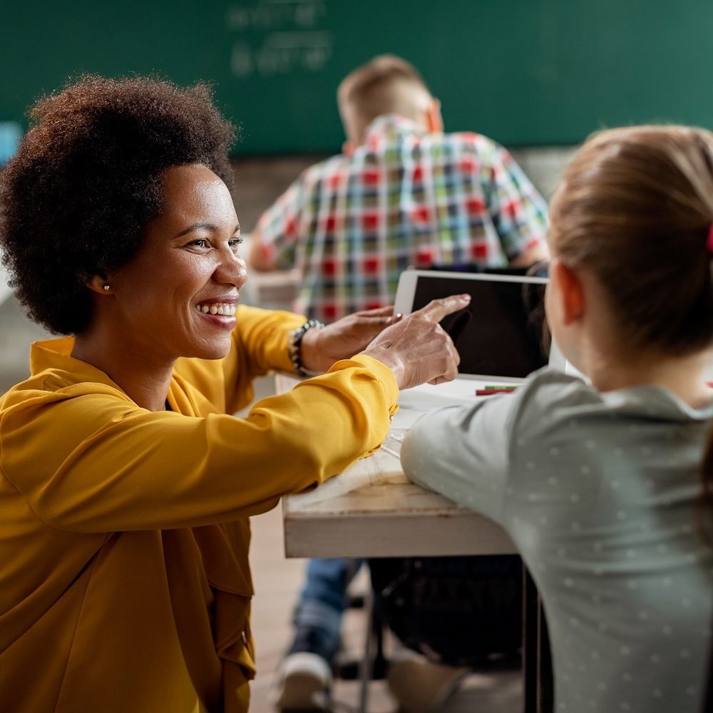 Happy African American teacher and elementary student using touchpad during a class in the classroom.